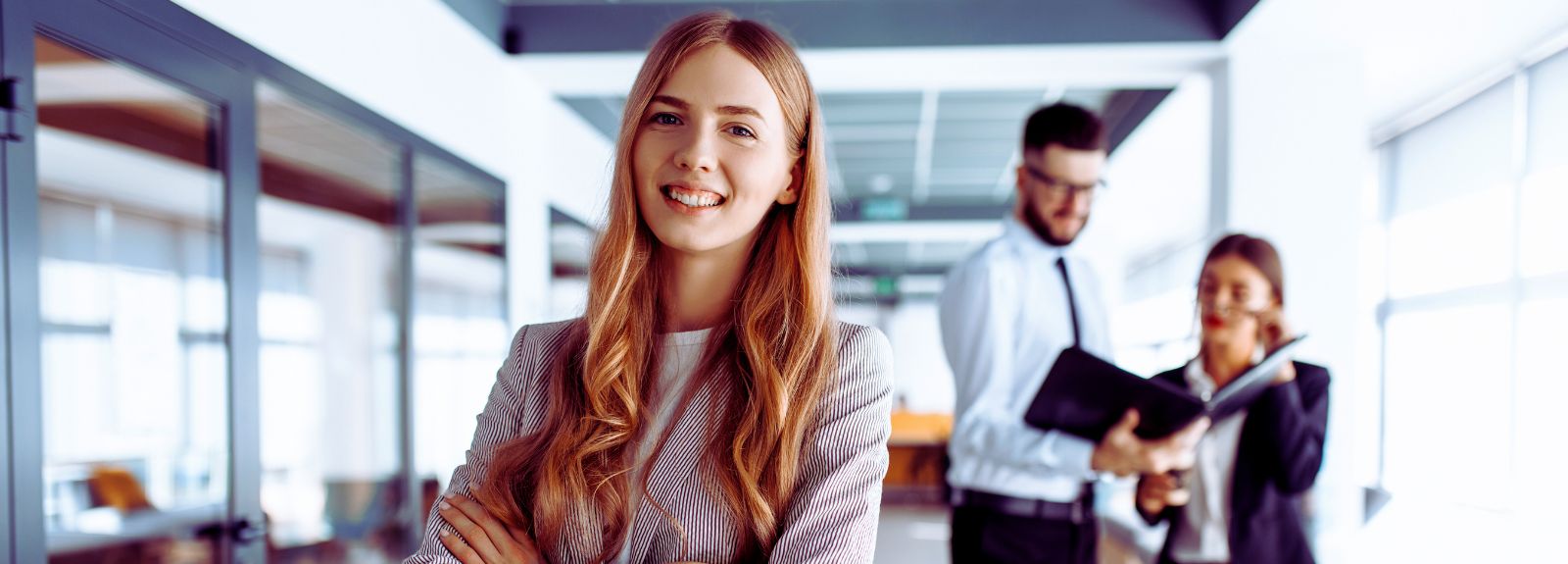 female standing in an office looking to camera, two colleagues talking in background